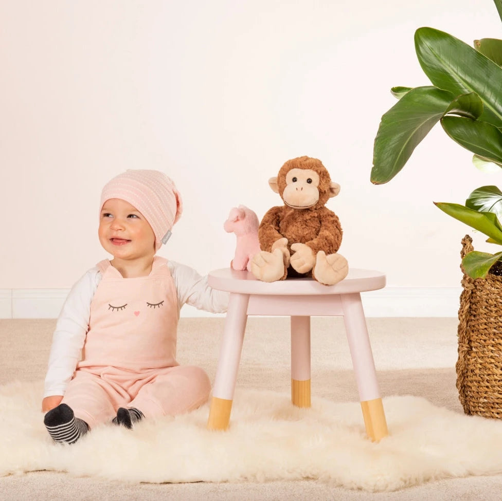 Baby in pink outfit with monkey plush toy on stool, plant in background