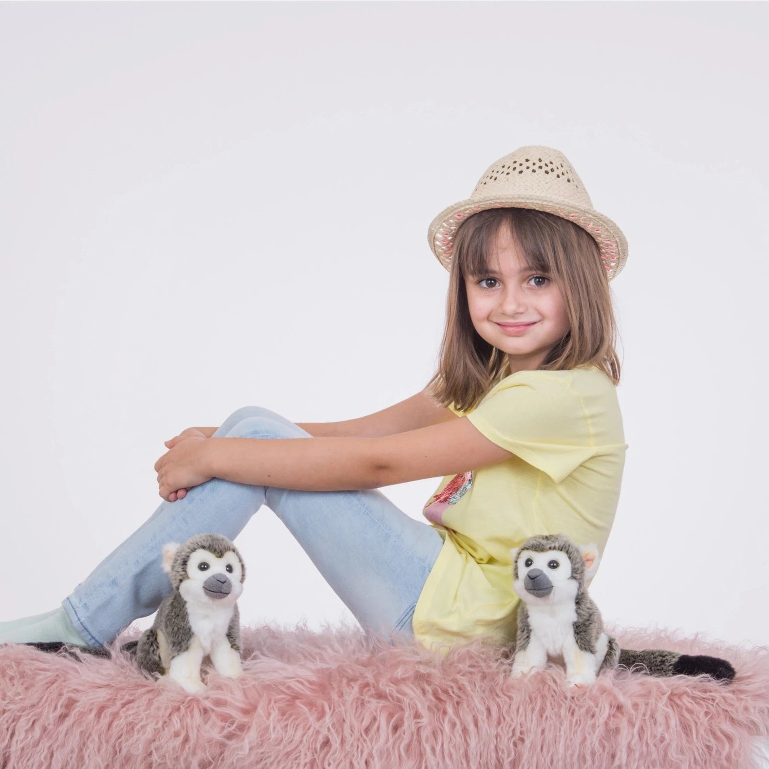 Young girl sitting on a pink fluffy surface with two stuffed animals, wearing a yellow shirt and light blue jeans.